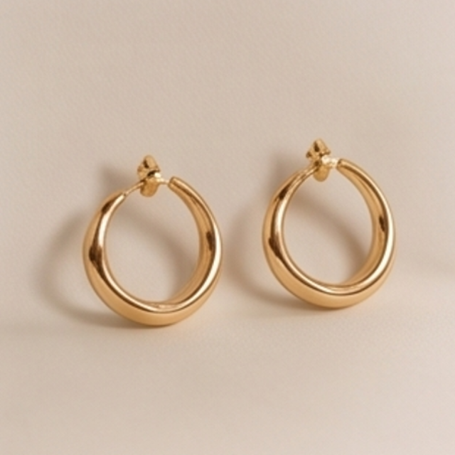A pair of polished gold-tone hoop earrings with a thick, rounded tubular design and post-back closures, displayed on a neutral beige background.
