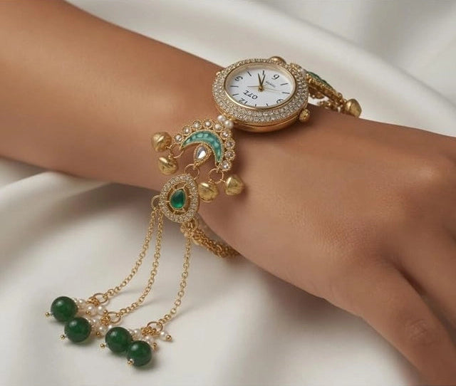 A gold-plated wristwatch with a decorative bracelet featuring green stone accents, crystal embellishments, and dangling chains, shown worn on a person's wrist against a white fabric background.